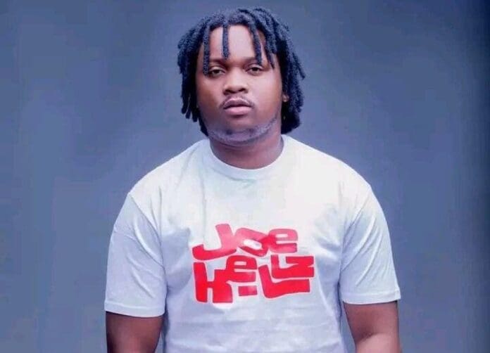 A man with short dreadlocks wearing a white t-shirt with "Joe Kellz" written in red stands against a plain blue background, representing the My Booth campaign.