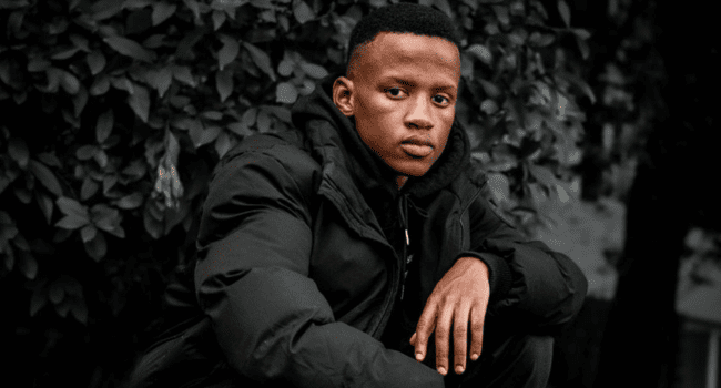 A young man in a black jacket sits outdoors in front of leafy bushes, looking directly at the camera with a neutral expression, embodying the cool confidence of Kane Keid—the rising star of African Rap and contender for Best Rapper in Africa.