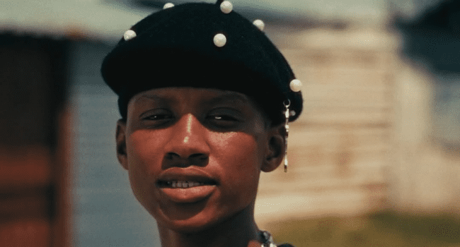A person wearing a black beret decorated with pearls stands outdoors, facing the camera, with a blurred background of buildings—channeling cool vibes reminiscent of Stogie T.