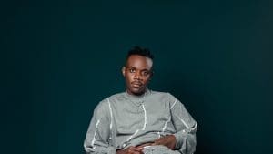 A person wearing a light gray, long-sleeved outfit sits against a dark teal background, looking directly at the camera with a neutral expression, channeling cool Afrobeat vibes inspired by Keltony.