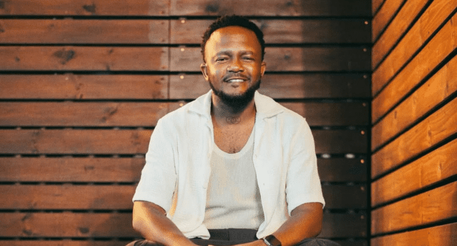 A man with short hair and a beard sits in front of a wooden wall, wearing a white shirt over a white tank top, looking like he's ready to join the next creative campaign with Mzansi Youth Choir or vibe out with Kwesta.