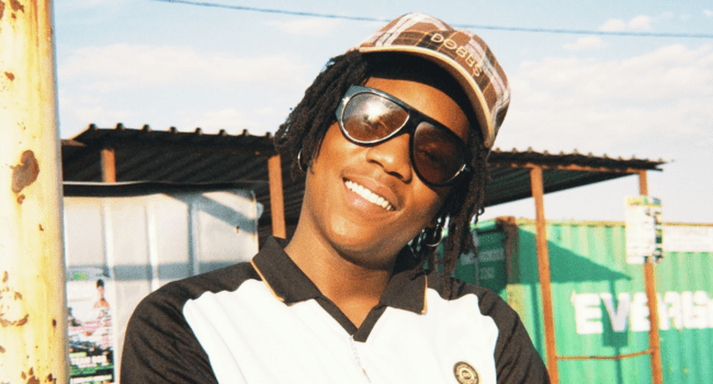 A person wearing sunglasses, a checked cap, and a black-and-white shirt stands outdoors, smiling with arms crossed in front of green shipping containers and a corrugated roof structure—channeling Nasty C’s confident debut album vibe.