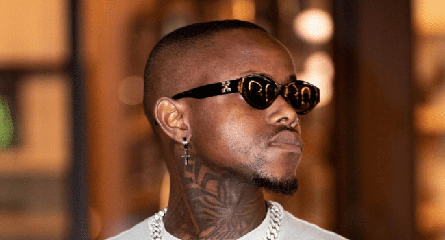 A man with closely cropped hair and a goatee wears dark sunglasses, a chain necklace, a cross earring, and has a neck tattoo—ready for the release of his upcoming single, “Loki,” this July.