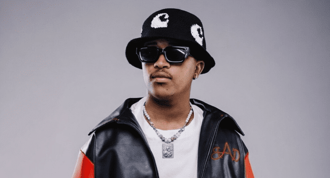 A person wearing a black bucket hat with a white pattern, black sunglasses, a necklace, and a black and red jacket—channeling that Lowfeye vibe—stands against a neutral background.