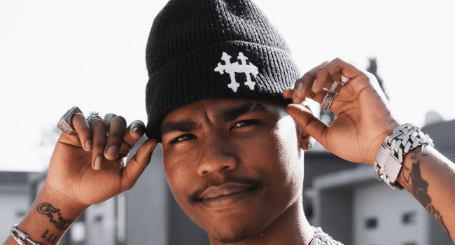 A person wearing a black beanie with a white symbol, silver jewelry, and black shirt, touching the edges of the beanie outdoors—channeling Lucasraps and Gemini Major vibes while waiting for new music to drop.