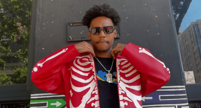 A person wearing sunglasses and a red jacket with a white skeleton design stands outdoors, holding the jacket open to reveal a black "Call Me" T-shirt with a colorful graphic.