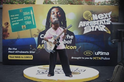 A person stands on a round platform playing guitar in front of a "Next Afrobeats Star" promotional backdrop at the Lagos venue, featuring an artist's image and event information.