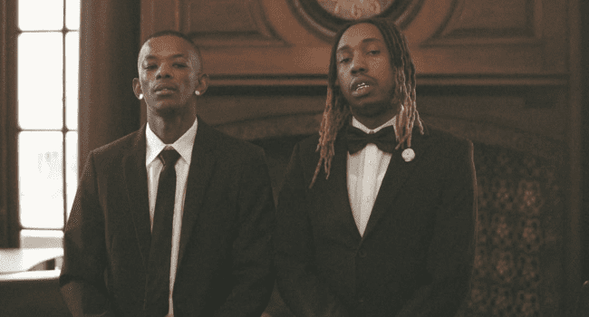Two men wearing suits, possibly Maglera Doe Boy and 25K, stand side by side indoors in front of a wooden mantle and window—perhaps hinting at an upcoming joint album—as they look directly at the camera.