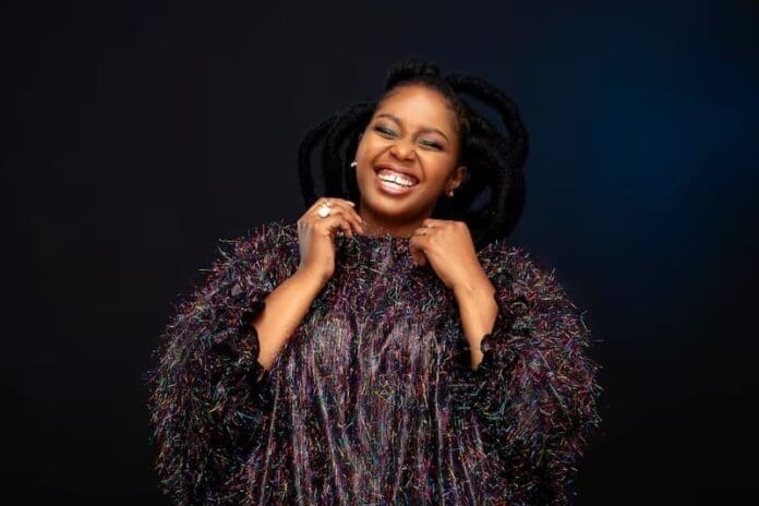 A woman with braided hair smiles broadly, holding the collar of her textured, dark, multicolored top against a plain background—capturing the excitement of the Maleh Album Launch on 28 March 2026 at SA Music Zone.