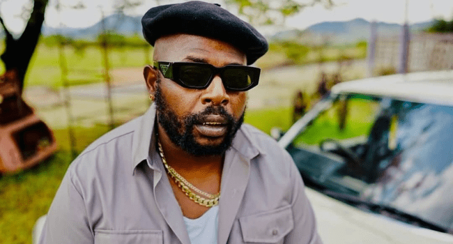 A man wearing a black beret, dark sunglasses, gold chains, and a gray shirt stands outdoors in front of a white car, capturing the spirit of SA Hip-Hop.