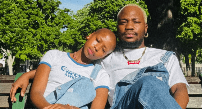 Two people wearing denim overalls and white shirts sit on a bench; one rests their head on the other’s shoulder. Sunlight filters through the trees behind them in this serene moment from the Fountains short film, set for a February release.