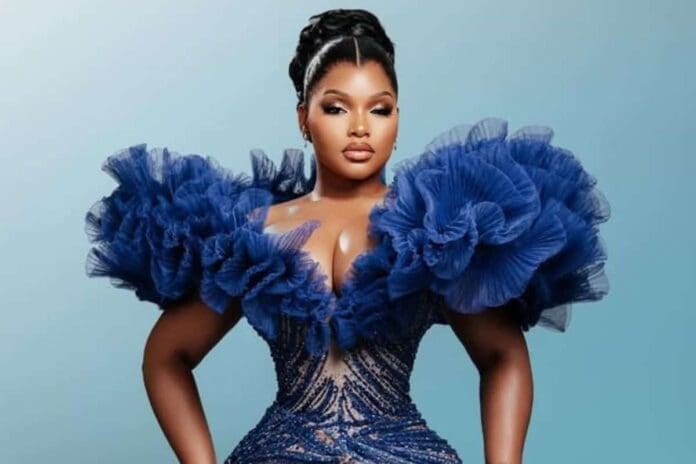 Woman wearing a blue, ruffled, off-shoulder gown with intricate detailing, standing against a simple blue background—Mawhoo shines with elegance in this male-dominated scene, echoing the bold spirit of Amapiano music.