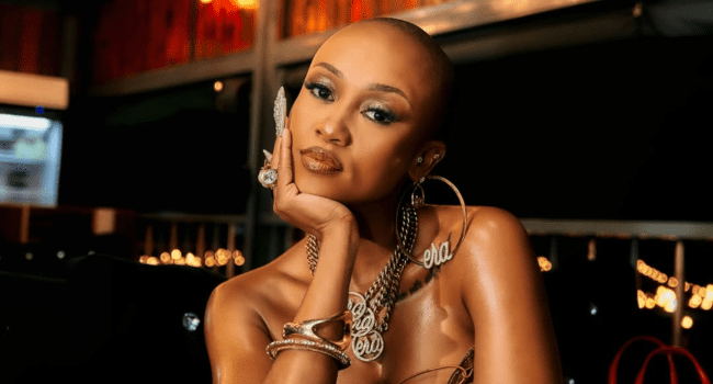 A woman with a shaved head, wearing a silver necklace and earrings, rests her chin on her hand while gazing at the camera in a dimly lit indoor setting, evoking the stylish vibe seen in Moozlie's new music visuals.