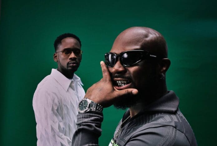 Two men pose in front of a green background; King Promise flashes his grill in sunglasses and a watch, while Mr Eazi stands further back in a white textured shirt.