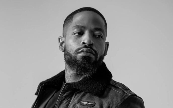 A man with a beard and short hair wearing a leather jacket looks to his left against a plain background, evoking the cool confidence of Prince Kaybee.