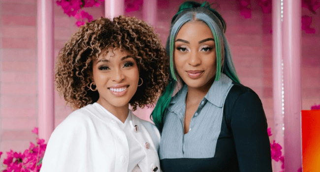 Two women smile side by side at a Naked Without Masterclass event. One has curly hair and a white outfit; the other, long hair with green highlights and a black and gray top. Pink flowers accent the background.