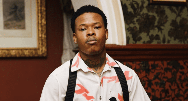 A man with short hair and visible neck tattoos stands indoors, wearing a white shirt with red patterns and black suspenders—a look reminiscent of Nasty C's rise in the music industry. Ornate wallpaper and framed art are in the background.