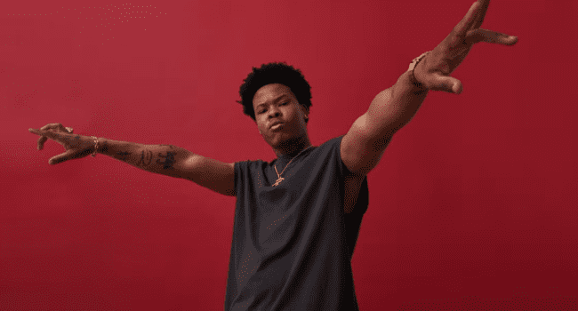 A person with short curly hair stands in front of a red background, wearing a sleeveless shirt, with both arms extended outward and a neutral expression, resembling Nasty C after he stopped going to church.