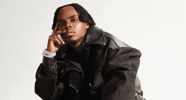 A person wearing a black textured jacket poses seated against a plain white background, channeling the reflective vibe of Ndu Browns' solo single, with one hand resting on their face and the other on their knee.