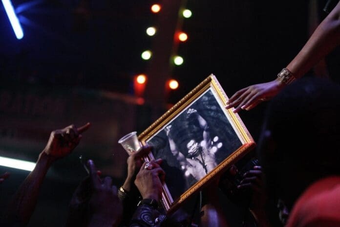 People in a crowd hold up a framed black-and-white photograph of Fela Kuti, the pioneer of African music, while reaching hands and a plastic cup toward it under colorful lights.