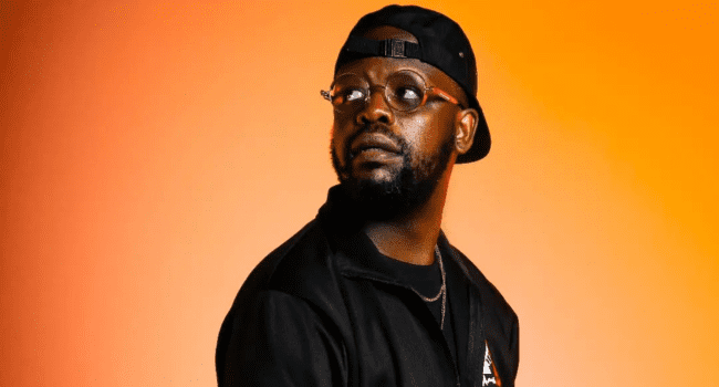 A man wearing glasses, a black cap, and a black jacket stands against an orange background, capturing the style of the New Generation SA Hip-Hop scene as he looks slightly to his left.