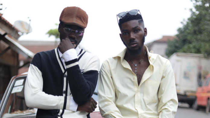 Two young men stand side by side outdoors; one wears a cap and glasses with a black-and-white cardigan, while the other, channeling Hiplife style, sports sunglasses on his head and an open-collared light shirt.