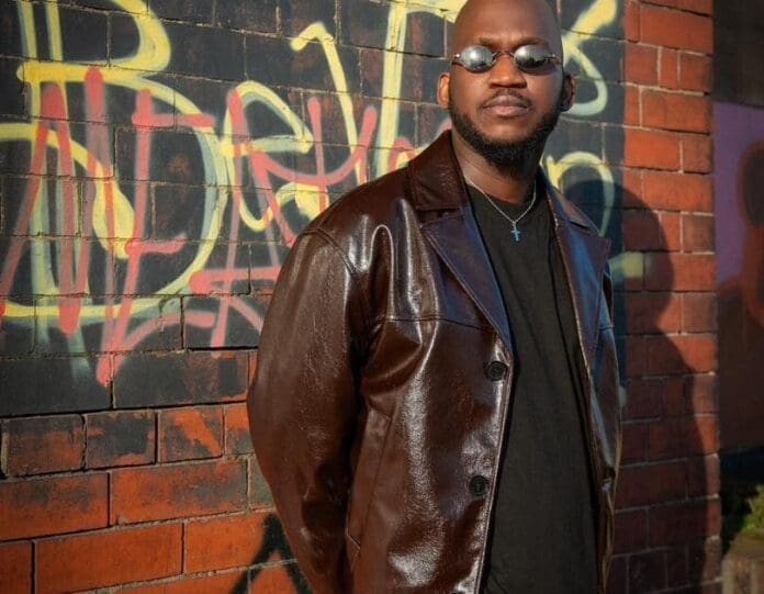 A man wearing sunglasses and a brown leather jacket stands in front of a brick wall with colorful graffiti, exuding peace of mind—an image captured by OfficiallyPiro.