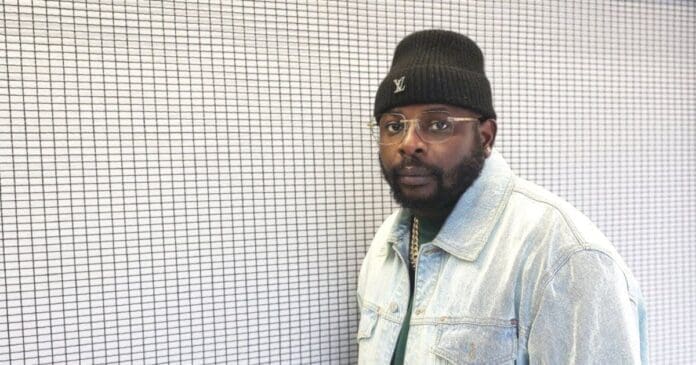 A man wearing a black beanie, glasses, and a light denim jacket stands in front of a white wall with a black grid pattern, looking ready to spin some amapiano songs like DJ Maphorisa.