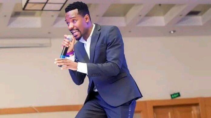 A man in a dark suit holds a microphone and appears to be speaking or singing on stage in a brightly lit indoor venue, reminiscent of Namadingo seizing his Grammy chances.