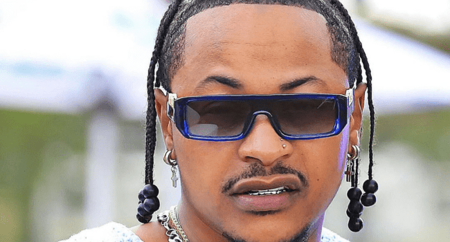 A person with braided hair, large sunglasses, earrings, and a nose piercing looks forward, reflecting on personal milestones like Priddy Ugly’s bold 2026 move.