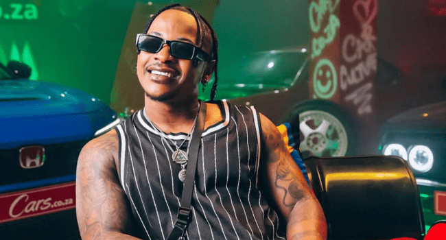 A person wearing sunglasses and a sleeveless striped shirt sits smiling in front of colorful cars with neon lights in the background, capturing the vibrant energy of a Priddy Ugly music video.