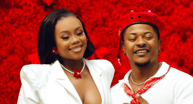 Two people dressed in coordinated white and red outfits stand smiling before a vibrant red floral background, radiating respect in marriage—a scene reminiscent of Bontle Modiselle's iconic style.
