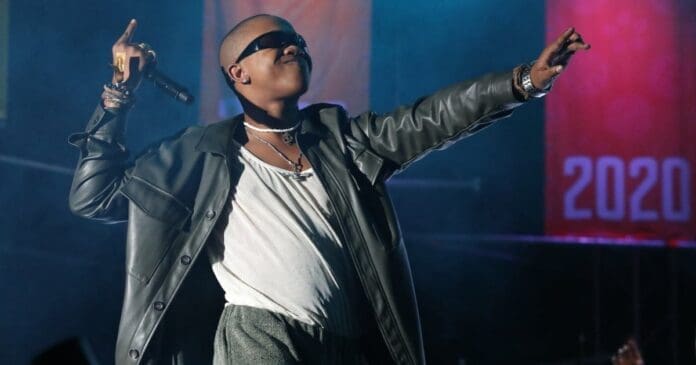 A performer, channeling Amapiano vibes in sunglasses and a leather jacket, holds a microphone and gestures with both arms on stage, with a "2020" banner in the background.