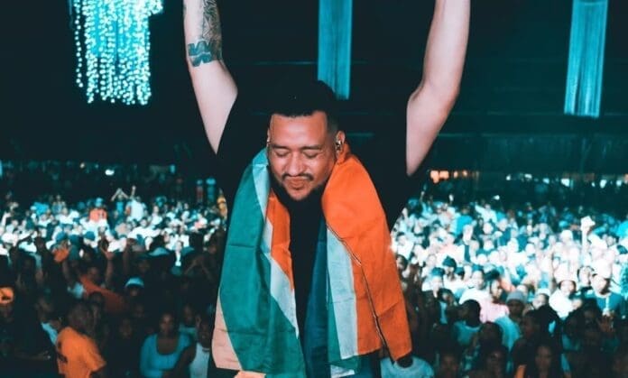 A man with arms raised stands on stage with a crowd in the background, draped in a flag, under hanging blue lights—capturing the spirit of SA Hip Hop.