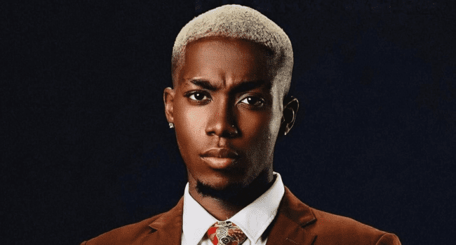 A man with short, platinum blonde hair wears a brown suit, white shirt, and patterned tie, posing against a dark background—channeling the confident style seen on Freestyle Fridays.