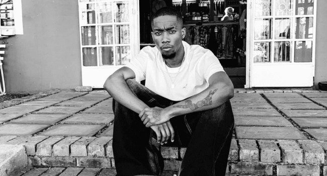 A man sits on a brick step in front of double doors, wearing a white t-shirt and dark pants, channeling Friday Freestyle energy as he looks directly at the camera.