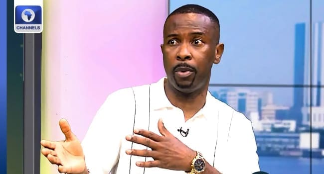A man in a white shirt gestures with his hands while speaking on a Channels Television show set, with a cityscape in the background, discussing topics like Afrobeats.