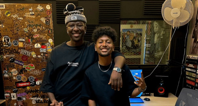 Two men, including producer Shaney Jay, stand together in a recording studio, smiling at the camera. One has his arm around the other. Studio equipment and a sticker-covered door are visible in the background.