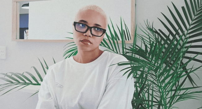 Person with short bleached hair and black glasses wearing a white shirt sits indoors near a green leafy plant, with a large mirror on the wall behind them—channeling Sammie Heavens’ cool vibe ahead of her forthcoming single, "My Man.