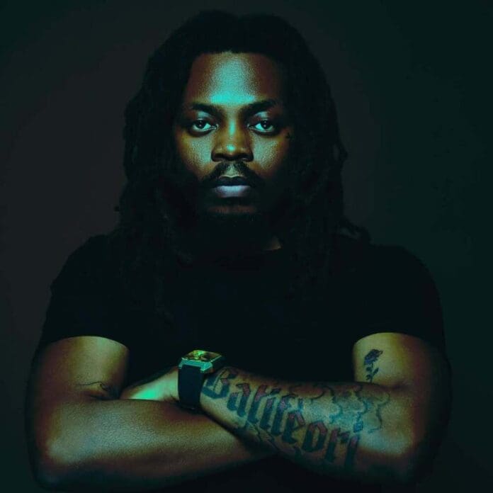 A man with long dreadlocks and a beard, wearing a black shirt, crosses his tattooed arms in front of his chest against a dark background, channeling the cool vibe of Afrobeats songs and the bold style seen in Olamide’s new album.