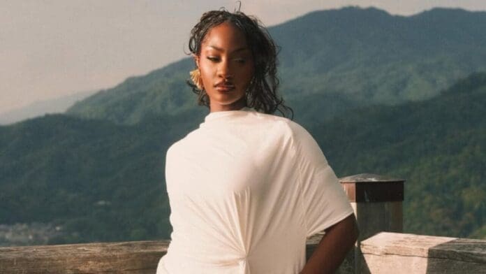 A woman in a white outfit stands outdoors in front of a wooden railing, with green mountains in the background, capturing Afrobeats vibes reminiscent of Tems’ iconic style.