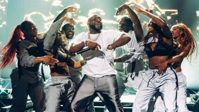 A performer in a white shirt dances energetically on stage to Afrobeats songs, surrounded by six backup dancers; dynamic lighting and stage effects are visible in the background.