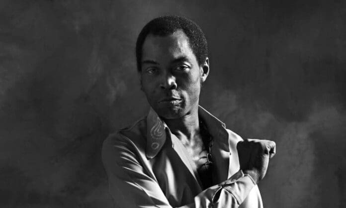 A man in a partially unbuttoned shirt stands against a plain, dark background, looking directly at the camera with one arm bent and hand resting on his shoulder, evoking the powerful presence of legendary African artist Fela Kuti.