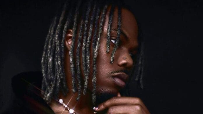 A person with shoulder-length, twisted hair and a nose piercing looks pensively to the side, resting their chin on their hand against a dark background, as if lost in thought while listening to Afrobeats songs.