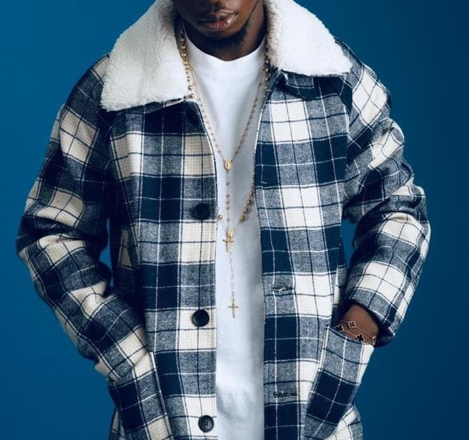 A person channels hip-hop style in a black and white checkered jacket with a white sherpa collar, a white shirt, and layered gold necklaces, standing confidently against a blue background.