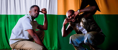 Two men posing in front of a large flag with green, white, orange, and black colors. Crouched down like Mr Eazi and King Promise in "That Way," they make expressive hand gestures near their faces.