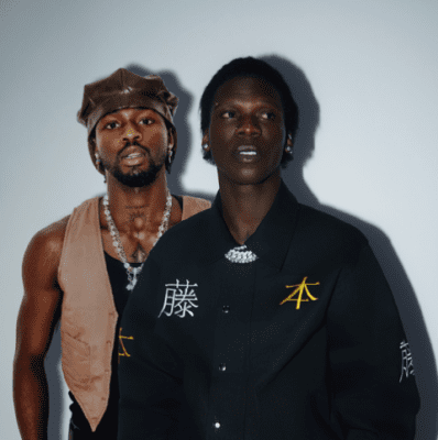 Two people stand side by side against a plain background; one wears a sleeveless vest and a leather cap, while the other sports a dark shirt with yellow Asian characters, channeling the bold style seen in Seyi Vibez's latest new single.