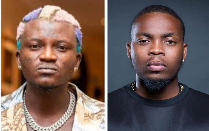 Two men facing the camera; the man on the left has dyed, multicolored hair and a chain necklace, while the man on the right sports a blacklist-worthy style with short, dark hair, a trimmed beard, and a black shirt.