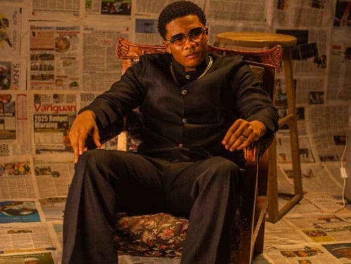 A man in dark formal attire and glasses sits on an armchair; the floor and walls, draped in newspaper pages, set a striking scene that feels like a backdrop for Tyler Vybe’s NEXT Wave in African Music.