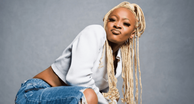 A person with long blonde braids, channeling Neo-Soul vibes in a white cropped sweater and ripped blue jeans, poses confidently in front of a gray background—ready to celebrate Scumie's new album.